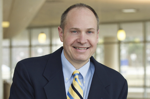 A portrait of Dr. Peter Henke. Dr. Henke wears a navy blue suit jacket with a light blue shirt and diagonally-striped blue and yellow tie.