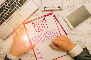 A clipboard with the message "Quit Smoking" is on a table top with a laptop and tablet