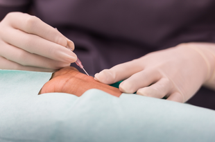 A health care provider wearing gloves and blue scrubs inserts a needle into a patient's wrist. 