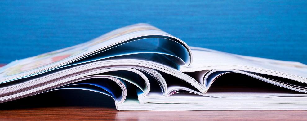 A stack of magazines lie open on a wooden table in front of a blue background.