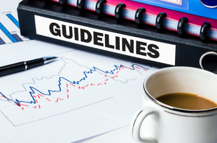 A binder labeled "guidelines" rests on a table on top of a line graph in red and blue. Also in the frame is a pen and a mug of coffee.