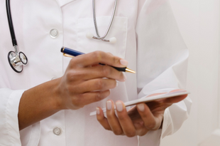 In a closeup, a Black, female physician wears a white coat and stethoscope. Her hands hold a pad of prescription paper and a pen, poised to write a prescription. 