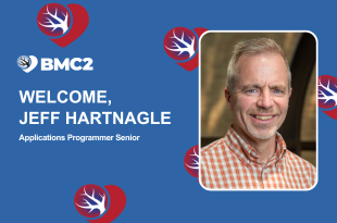 A white version of the BMC2 logo and the words, Welcome, Jeff Hartnagle Applications Programmer Senior are on a medium blue background along with red, blue, and white BMC2 hearts scattered around. A portrait of Jeff Hartnagle is to the right. Jeff has light-colored hair and facial hair, is smiling, and is wearing an orange and white plaid button-down shirt.