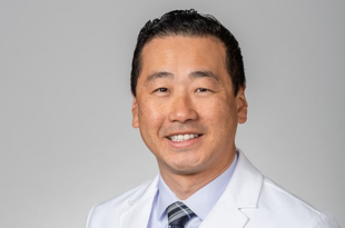 Dr. Ryan Kim has short dark hair and is wearing a white coat over a white shirt and dark tie with a light-colored plaid design.