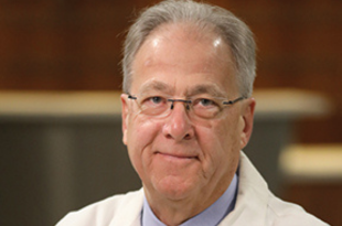 Dr. Ronald Miller smiles while wearing a light blue button-down shirt, white coat, and glasses.