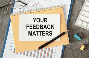 A piece of paper stating "Your Feedback Matters" is on a table top surrounded by a pair of glasses, a sheet of paper with graphs, a notebook, binder clips, and a keyboard.