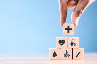 A stack of blocks with images of a medical cross, heart with an EKG, pills, a blood droplet, a patient in a wheelchair, and a needle and syringe are being stacked on a light-colored table on a blue background.