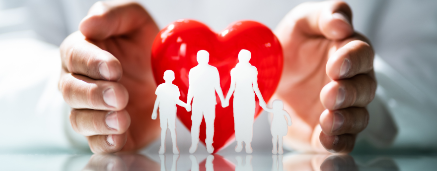 A closeup of a doctor's hands flanking a red heart and a white paper cut out of a family with a man, woman, and two children.