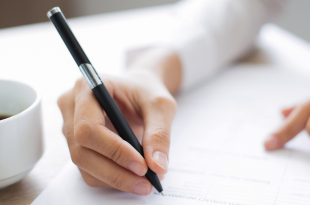 A female hand holds a black pen and signs paperwork. A cup of coffee in a white mug is to her right.