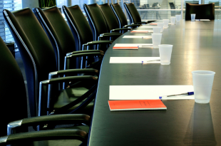A board room table is set for a meeting with cups, paper, pens, and red folders.