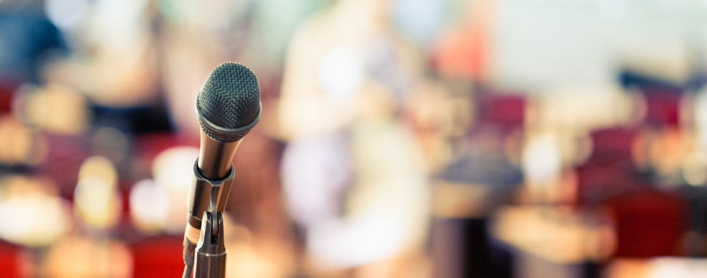 A microphone is in the foreground with an audience out of focus in the background.