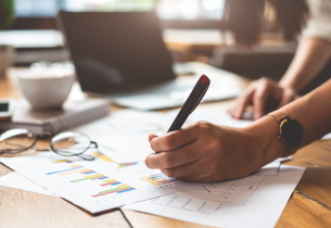 A person makes notes on reports that are scattered on a desk.