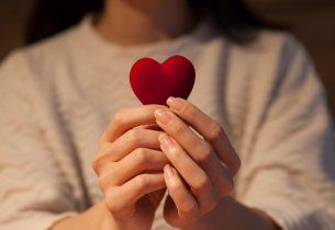 A woman with dark hair and wearing a white sweater holds a red heart.