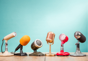 A row of microphones in various colors, shapes, and sizes sit on a table in front of a light blue background.