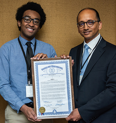 BMC2 Program Director, Dr. Hitinder Gurm, has short dark hair, dark-rimmed glasses, a dark suit coat, and white button down shirt with a medium-blue tie. He holds the framed tribute with Legislative Assistant, Zac Ozormoor who wears a medium-blue button down shirt and a black tie and has dark curly hair and dark-rimmed glasses.